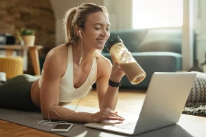 young-happy-sportswoman-drinking-smoothie-while-relaxing-floor-surfing-net-laptop